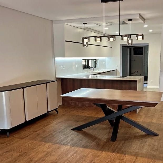 Espaço aberto com mesa de refeições em madeira com pés pretos cruzados, aparador curvo e cozinha ao fundo